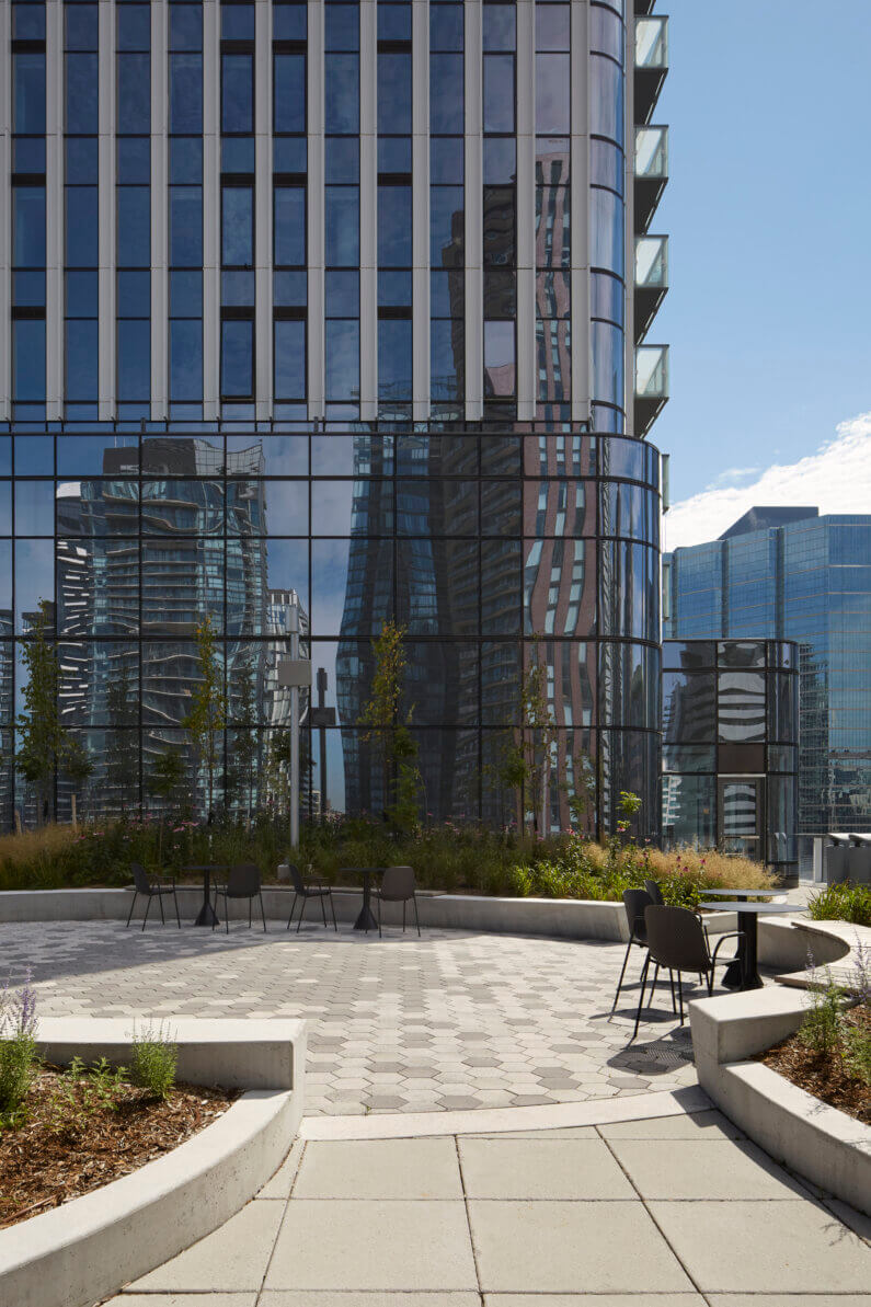 Outdoor seating area with hexagonal paving, surrounded by landscaping and reflecting glass buildings in the background. Clear sky overhead.