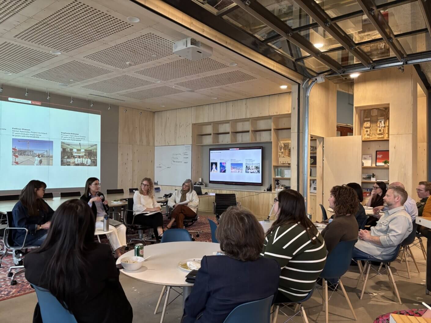 Panel discussion with multiple speakers in an architecturally designed space, featuring a projector screen and attentive audience members.