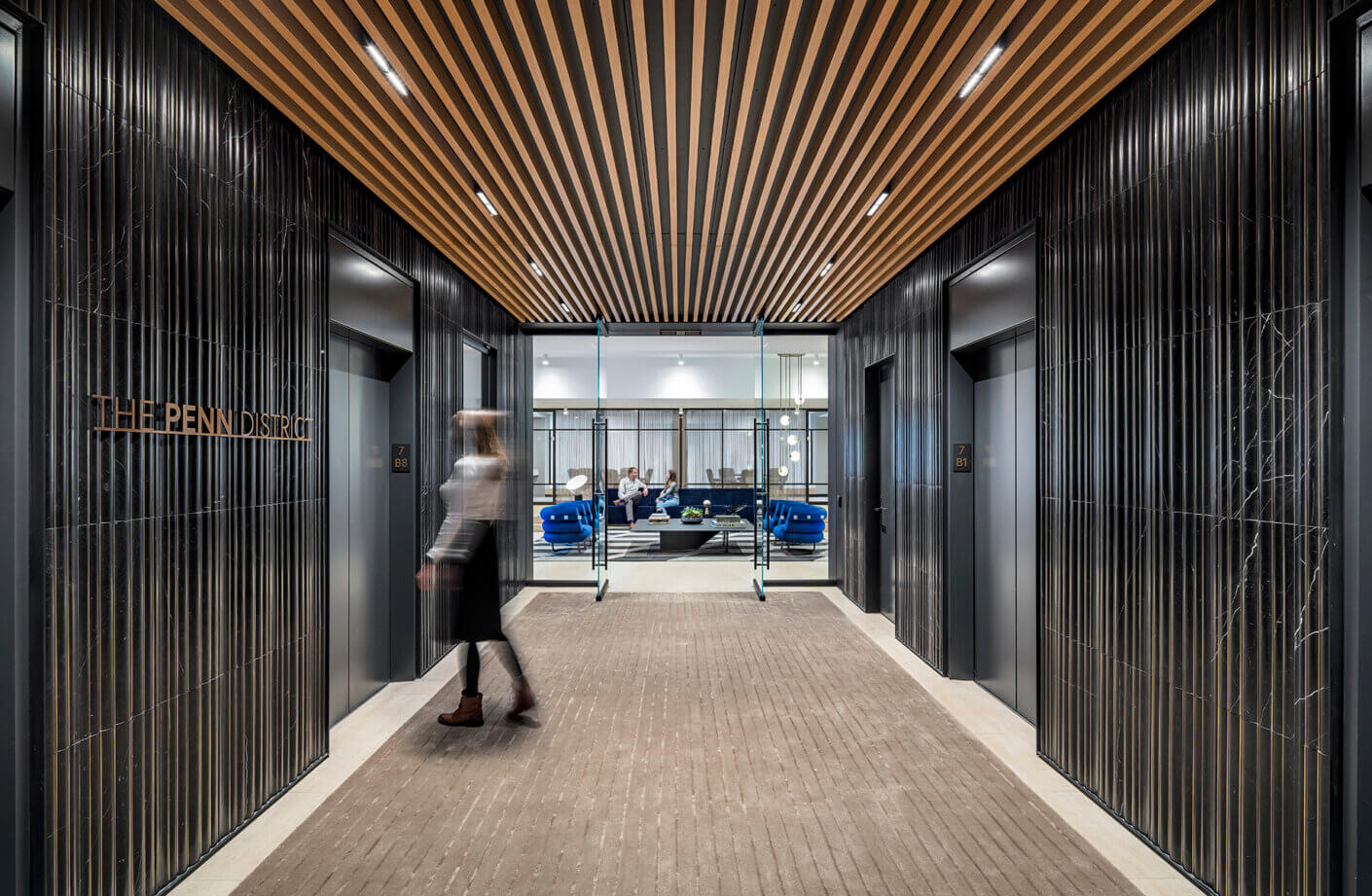 Elevator lobby at The Penn District featuring textured walls, wooden ceiling, and seating area visible through glass doors. Person walking by.