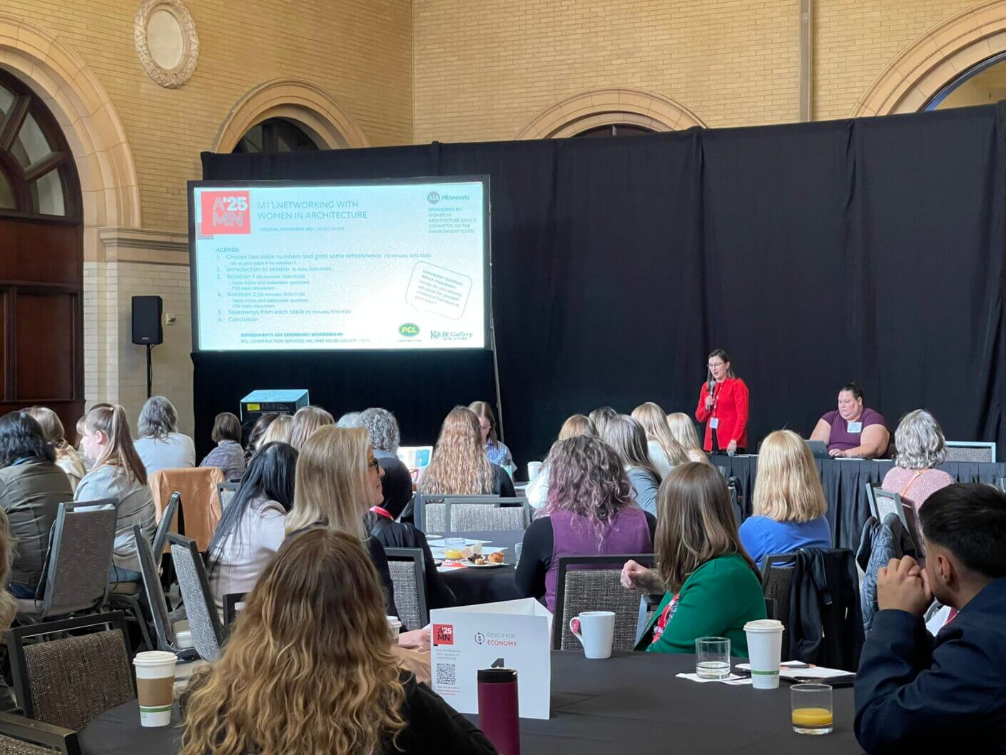Women in architecture networking event featuring speakers and attendees engaged in discussion at a conference. Agenda visible on screen.