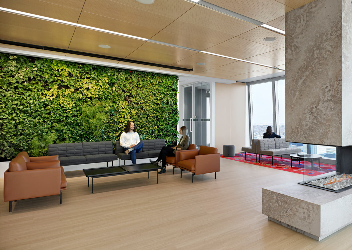 Office lounge featuring green wall, seating areas with brown and gray sofas, and large windows. People engaged in conversation and relaxation.