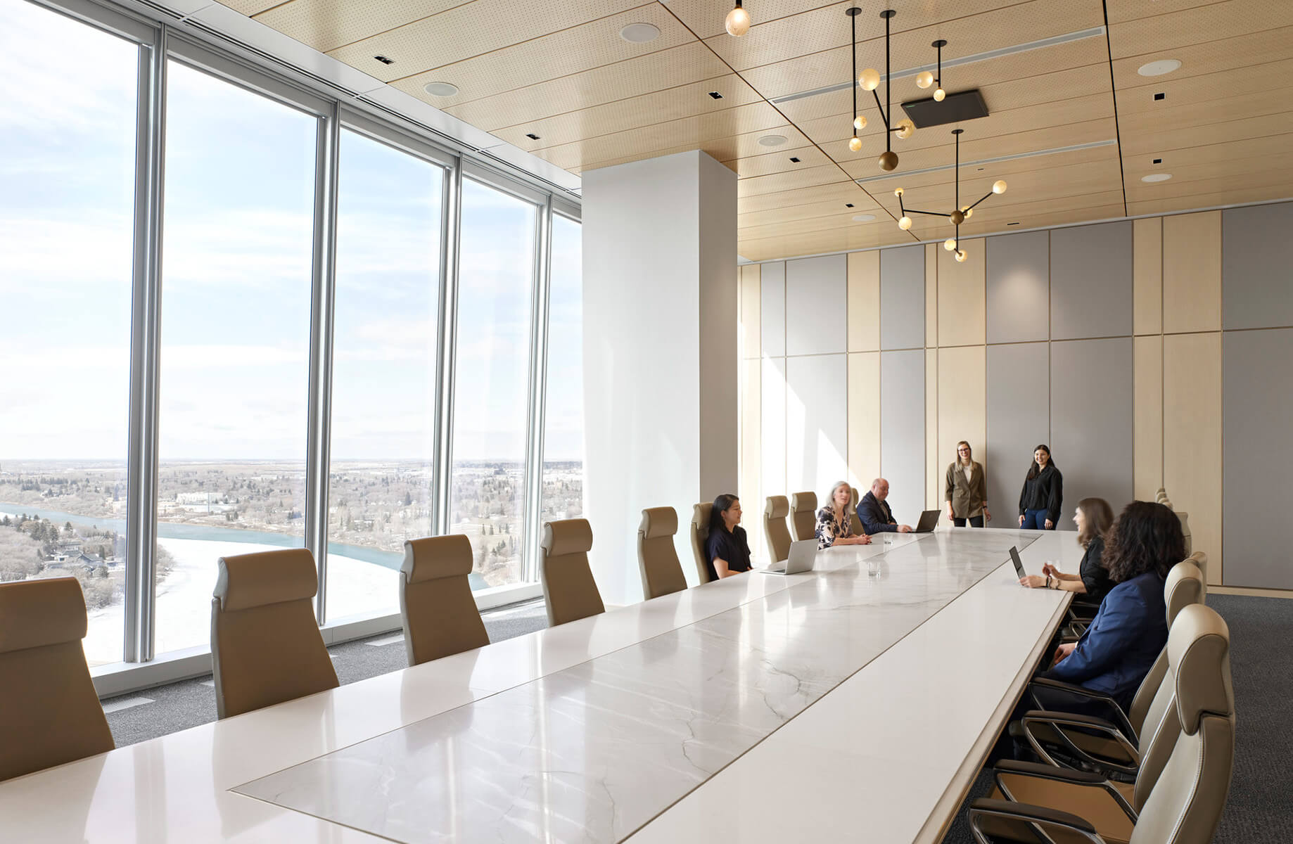Conference room with a long table, large windows, and participants engaging in discussion. Views of a river and landscape visible outside.