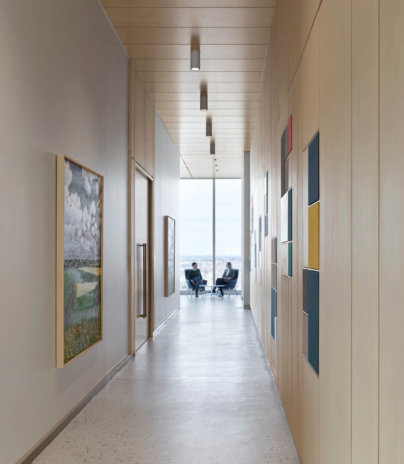 Hallway with wood paneling, artwork on walls, and natural light. Two people seated at the end near large window, creating a workspace atmosphere.
