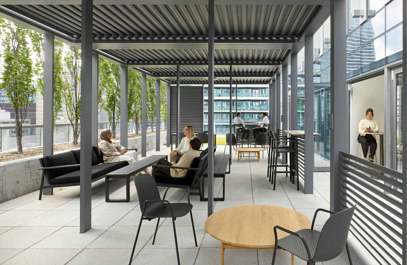 Outdoor workspace featuring seating areas with trees, a table, and people engaged in discussions and working on devices.
