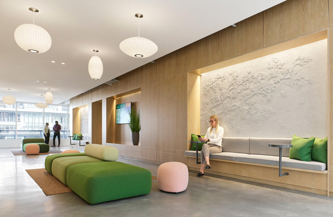 Two people converse in a spacious foyer, while a seated woman uses a tablet. The room features unique seating and a textured wall design.