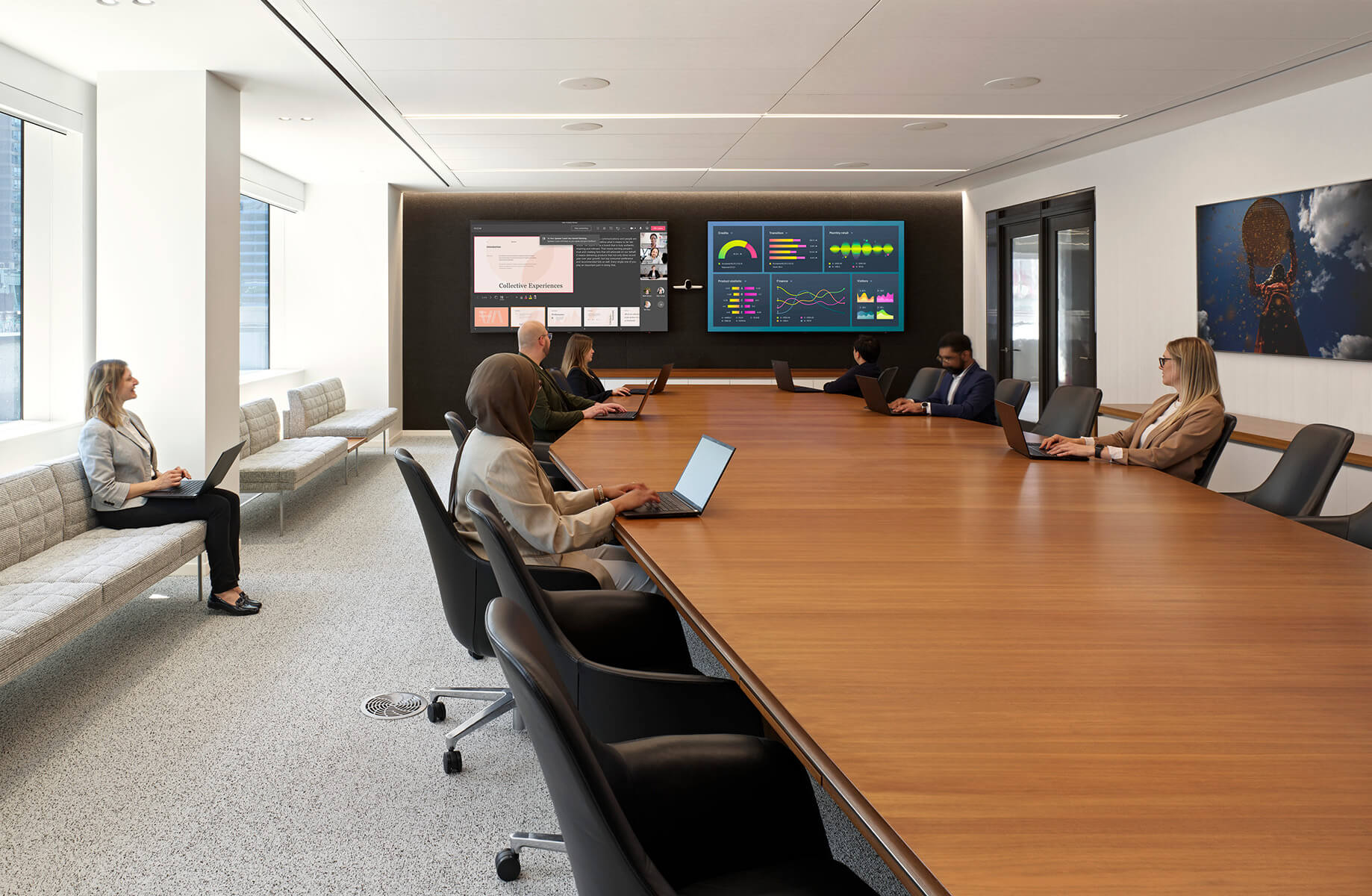 A conference room filled with seven professionals at a long table, engaged with laptops and presentations displayed on screens.