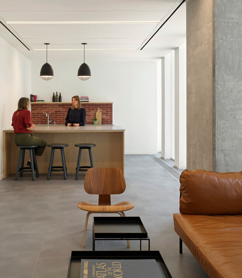 Two women converse at a kitchen island with wooden elements, black stools, and pendant lights. A brown leather sofa and wooden chair are visible in the foreground.