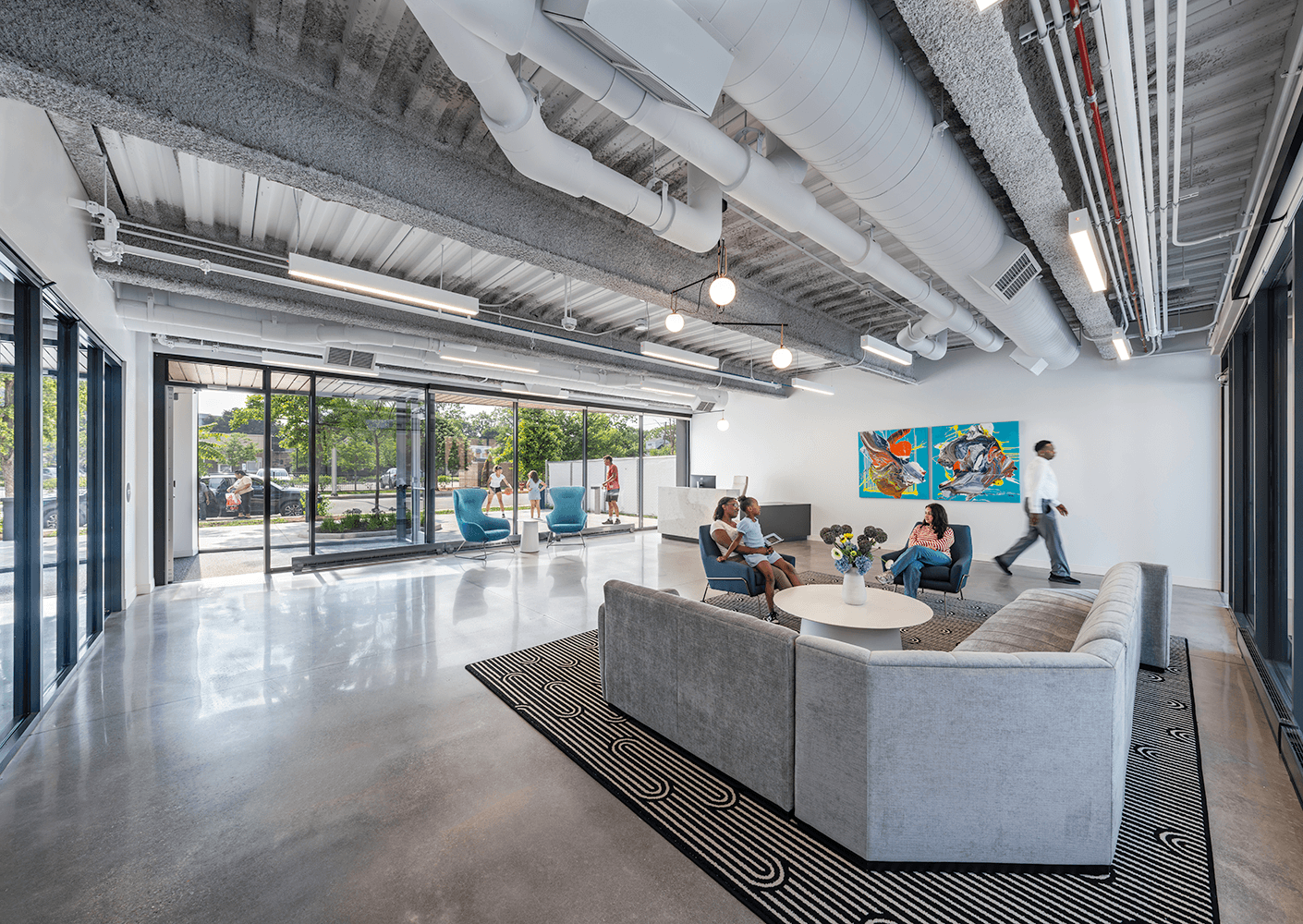 Waiting area with a gray sectional sofa, colorful artwork on the wall, and glass windows. Two people are seated while one walks by.
