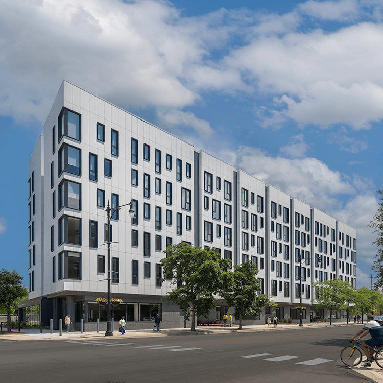 Urban residential building with multiple stories, large windows, and ground-level greenery, set against a clear blue sky.