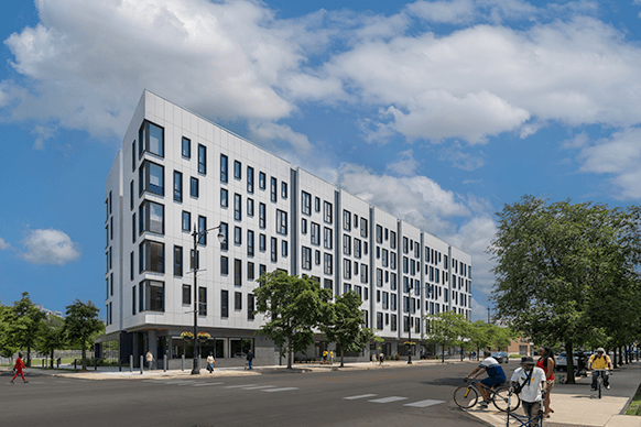 New residential building at a street intersection, featuring contemporary design and landscaping, with pedestrians and cyclists nearby.
