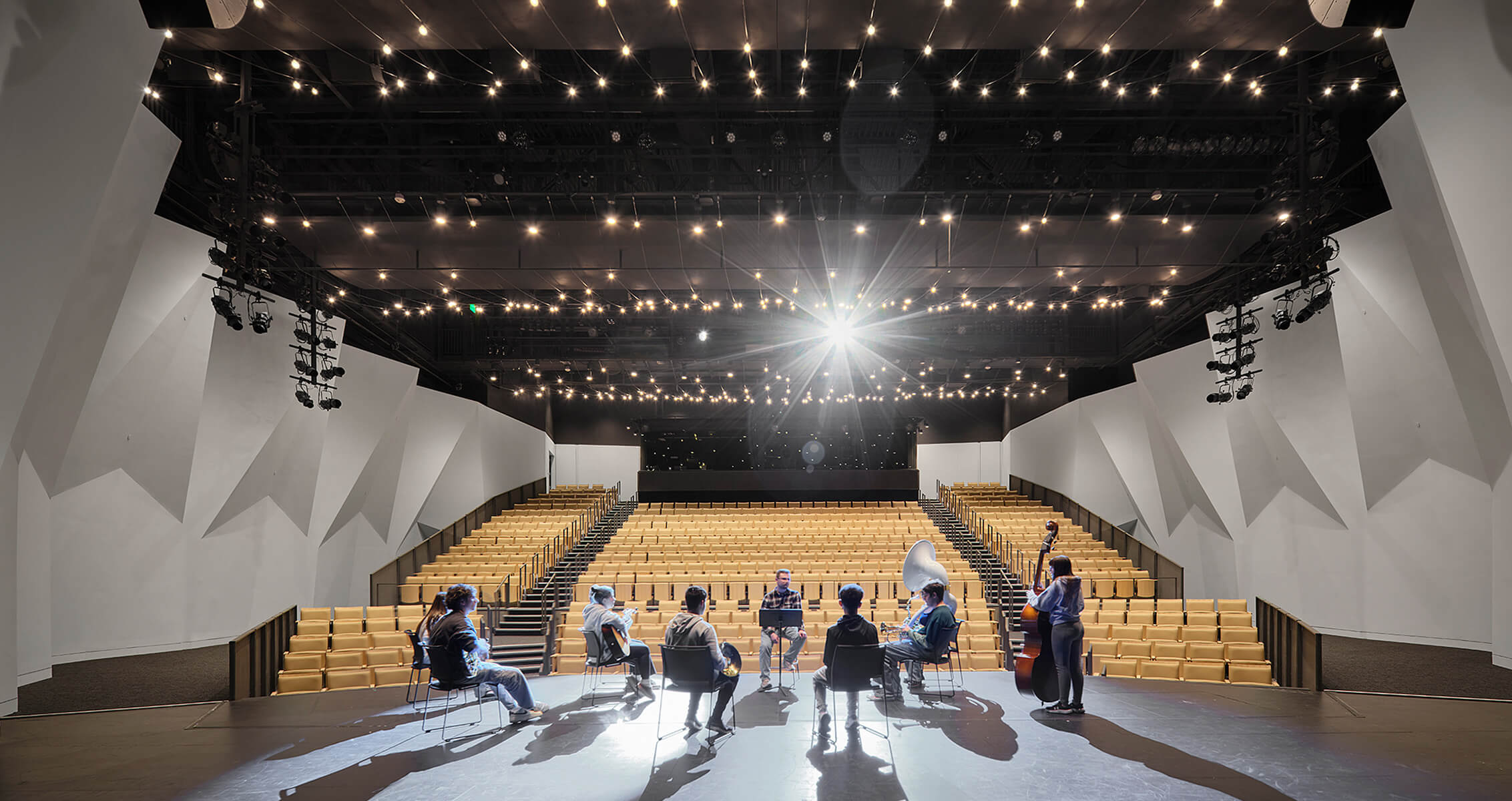 Rehearsal in a spacious auditorium with yellow seating, featuring musicians and bright stage lighting, set against geometric wall design.