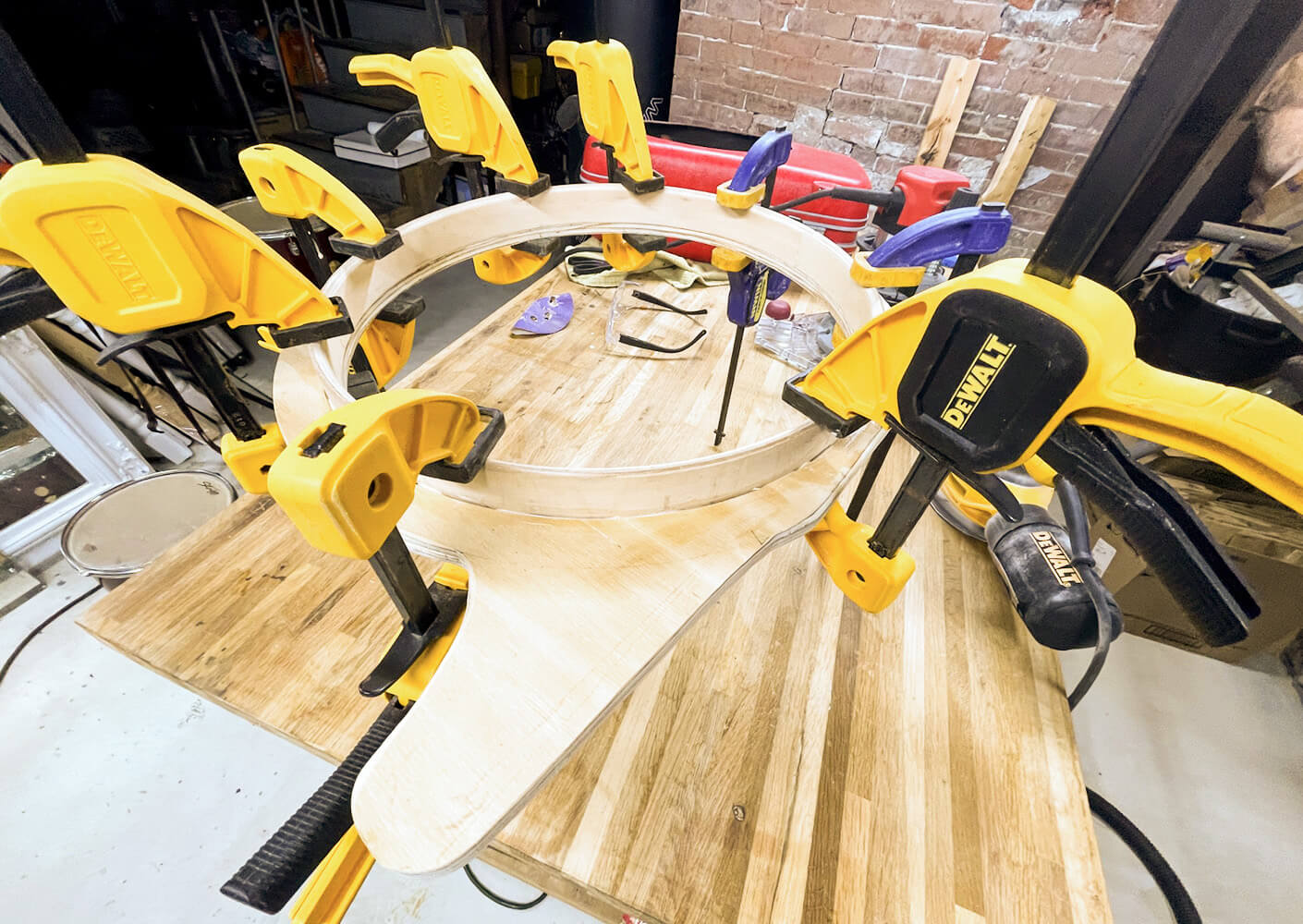 Clamps securing a wooden circular piece on a workbench in a workshop, with tools and materials in the background.