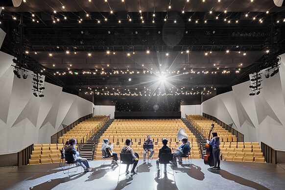 A group of musicians rehearses in an auditorium with tiered seating and overhead lights, showcasing a spacious performance area. 