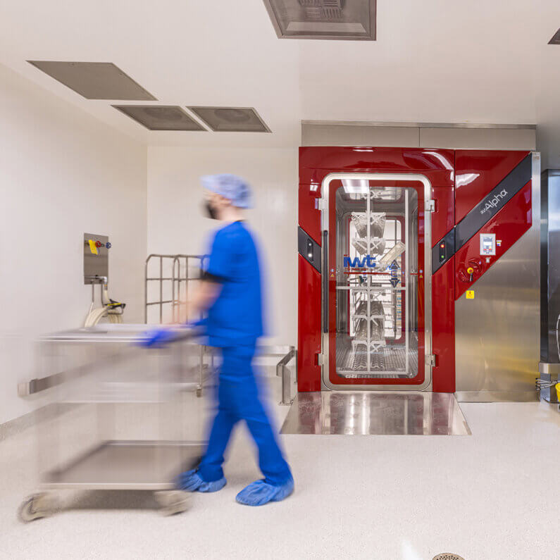 Lab worker in blue scrubs pushes a cart past an advanced red cage wash unit in a cleanroom environment.