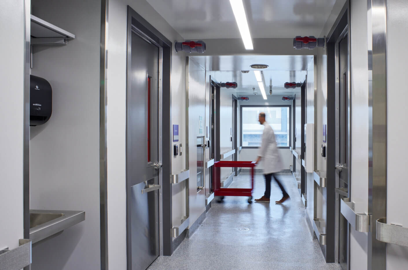 A healthcare professional in a lab coat walks through a clinical corridor, pushing a red cart past patient rooms and safety equipment.