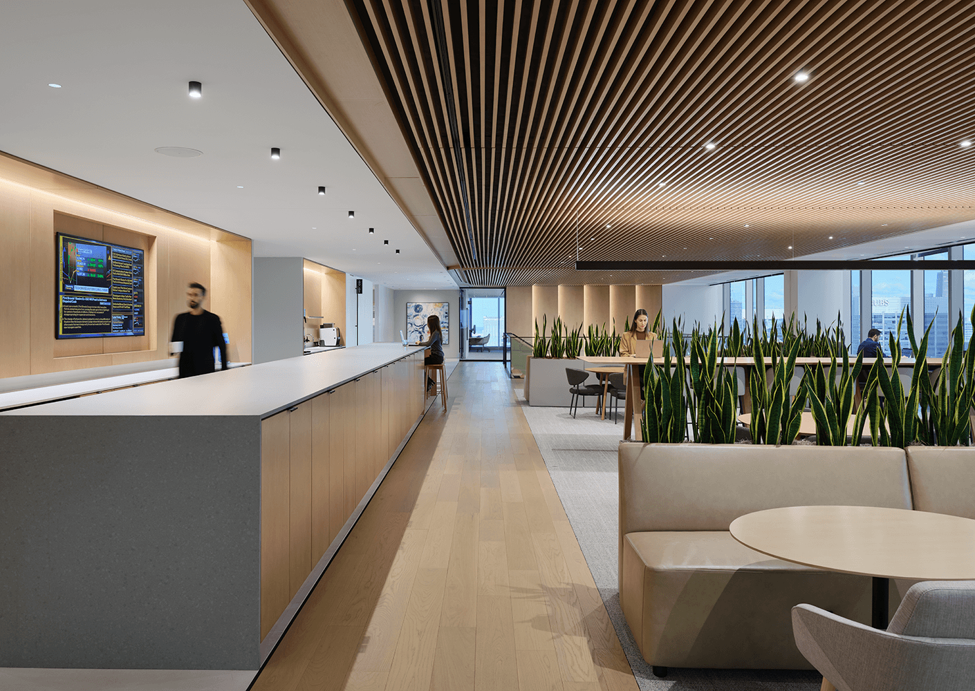 Interior view of a corporate space featuring a reception area, seating, greenery, and workstations, with wood paneling and natural light.
