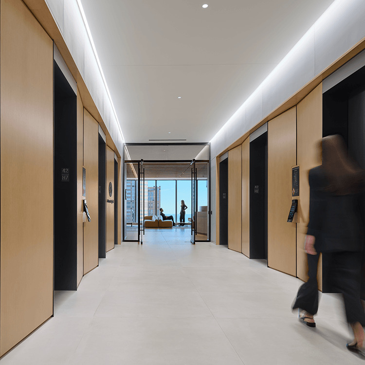 Elevator lobby featuring wooden paneling, large windows, and people interacting, with modern lighting illuminating the space.
