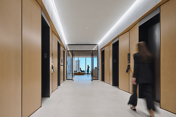 Elevator lobby with wooden paneling, spacious design, and large windows showcasing a city view. A person walks past the elevators.