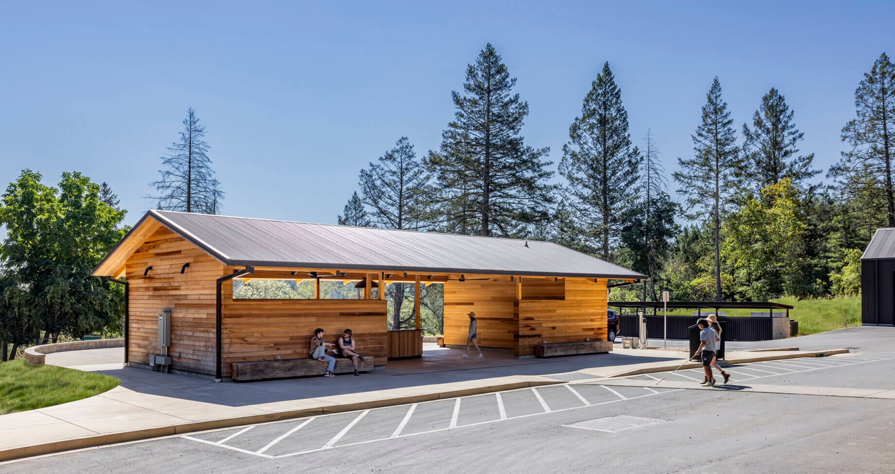 Wooden building with a metal roof surrounded by trees, featuring outdoor seating and people walking on a paved area.