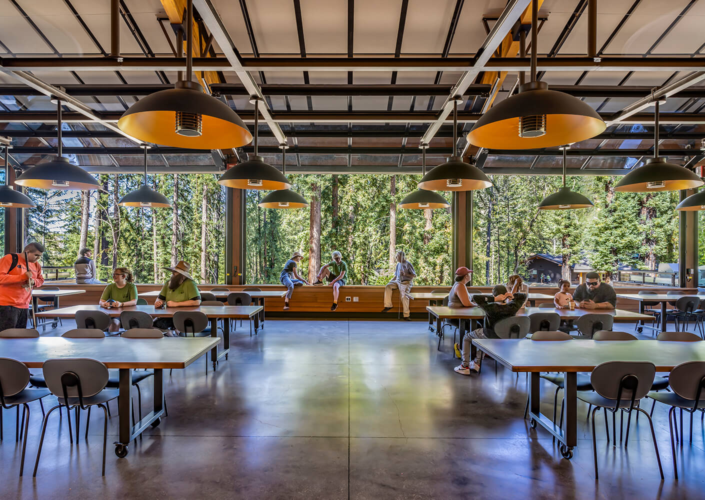 Interior view of a spacious dining area with tables, chairs, and large windows overlooking trees, featuring patrons engaging in conversation and activities.