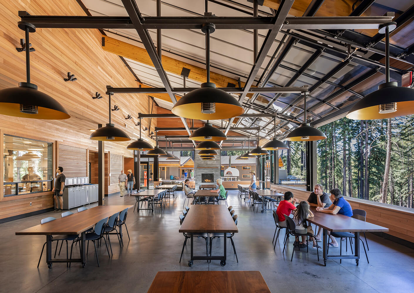 Interior view of a spacious dining area with wooden walls, pendant lighting, and tables, surrounded by large windows overlooking trees.
