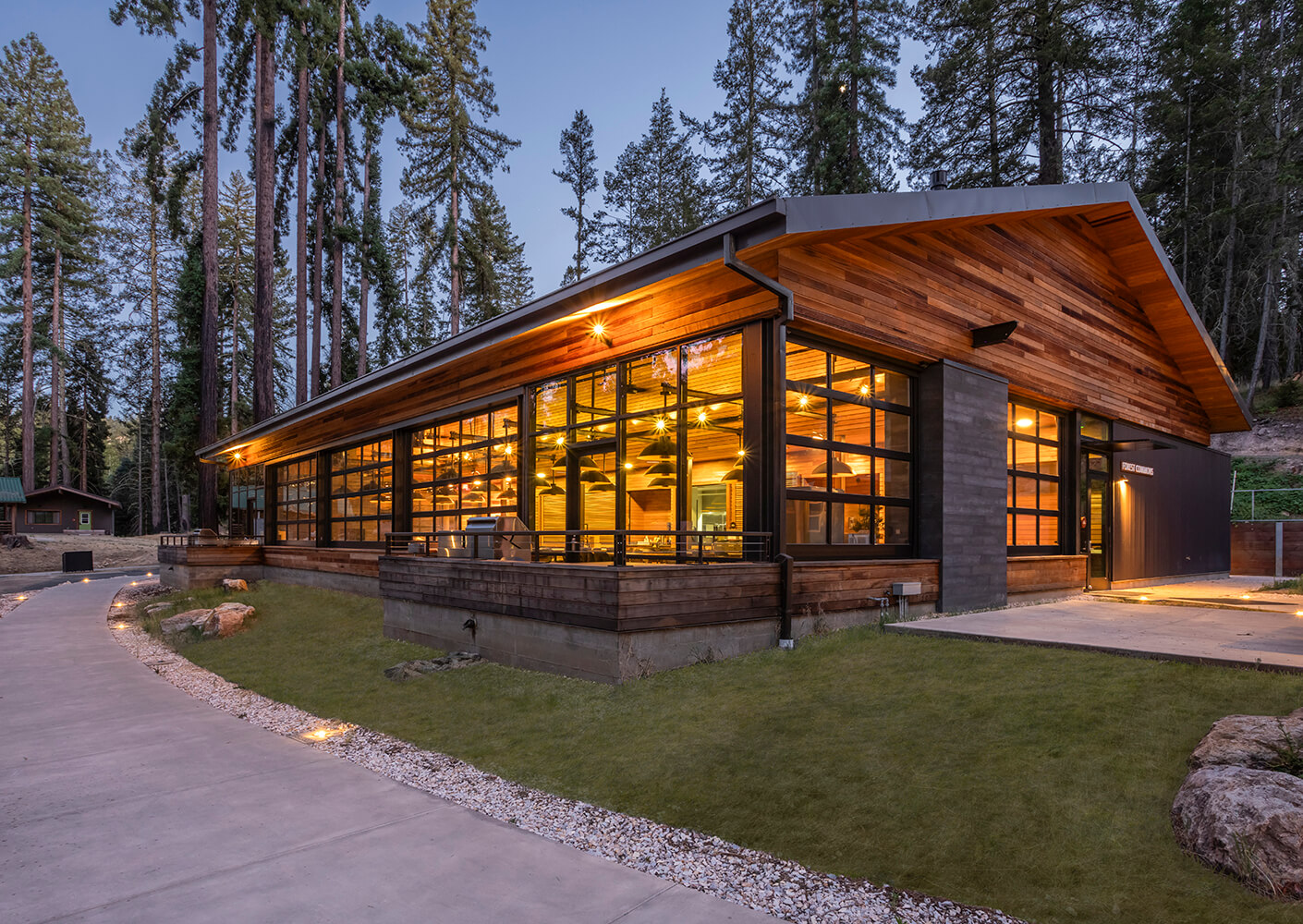 Architectural view of a cabin with large windows, surrounded by tall trees. Pathway leads to a well-lit entrance with a covered deck.