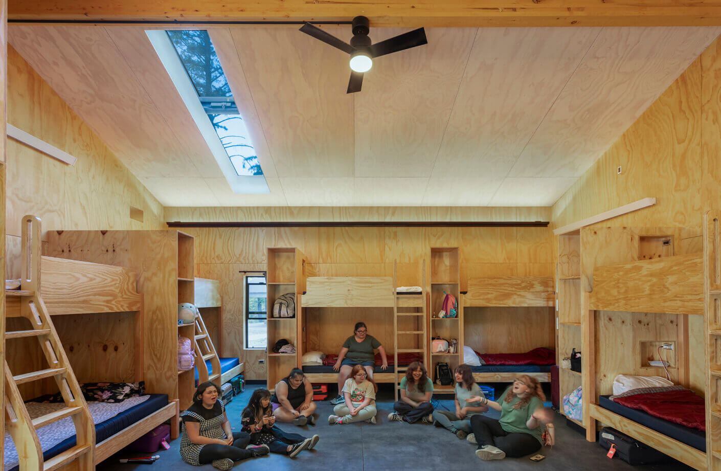 Group of people sitting on the floor of a room with wooden bunk beds, large windows, and a ceiling fan. Natural light illuminates the space.
