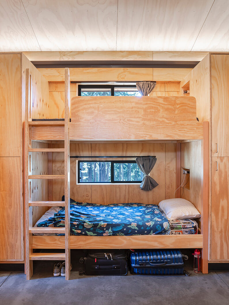 Bunk bed design in a compact space with wooden walls, featuring bedding, storage, and windows with simple curtains.