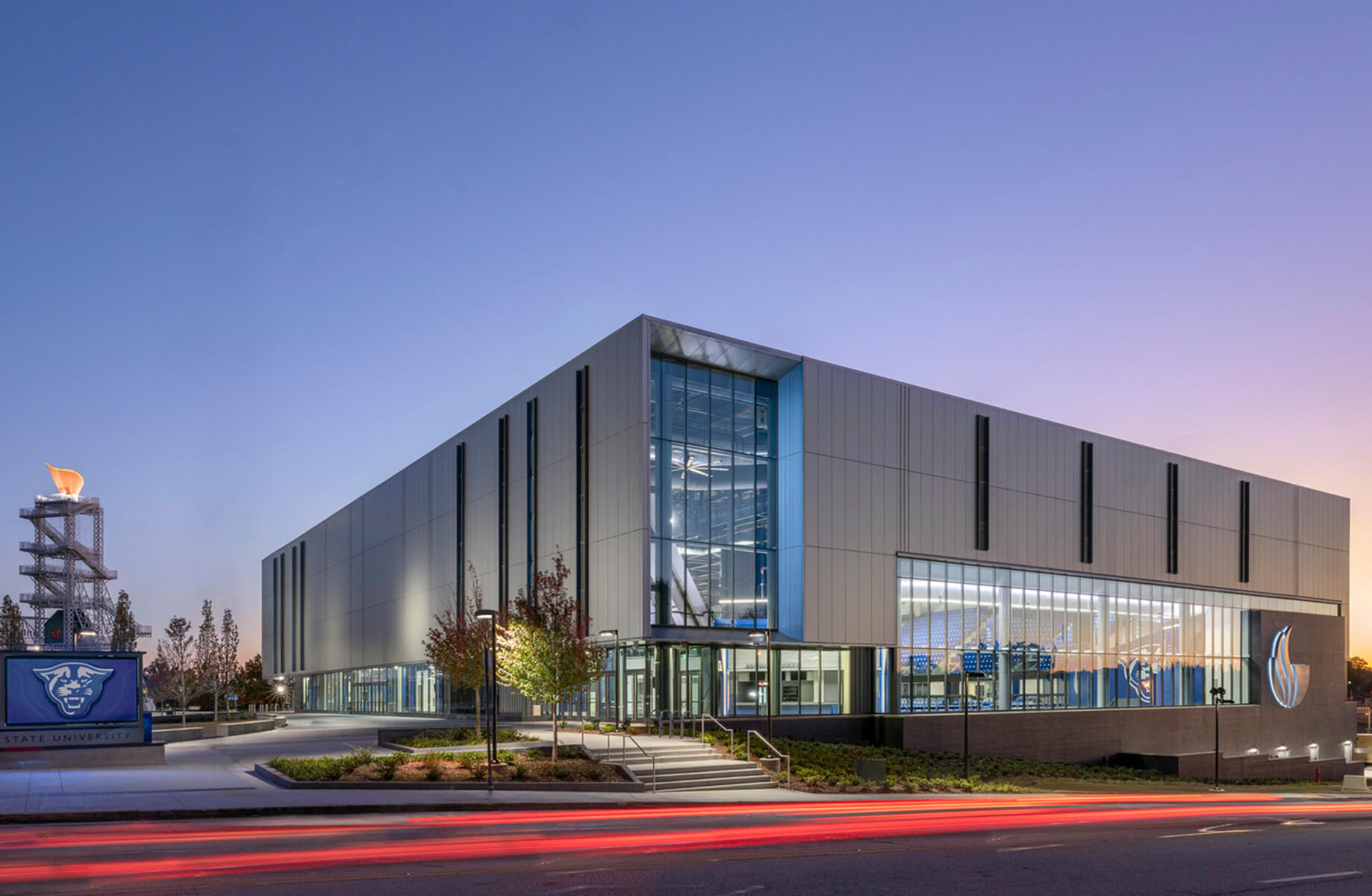 Architectural view of a university facility showcasing large glass windows and landscaping, with a prominent school logo and athletic flame sculpture.