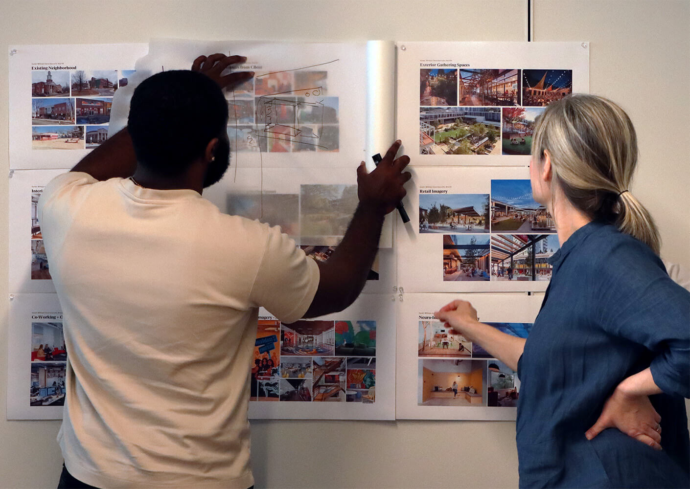 Two individuals review architectural design boards, discussing various project images and layouts for community spaces and retail designs.