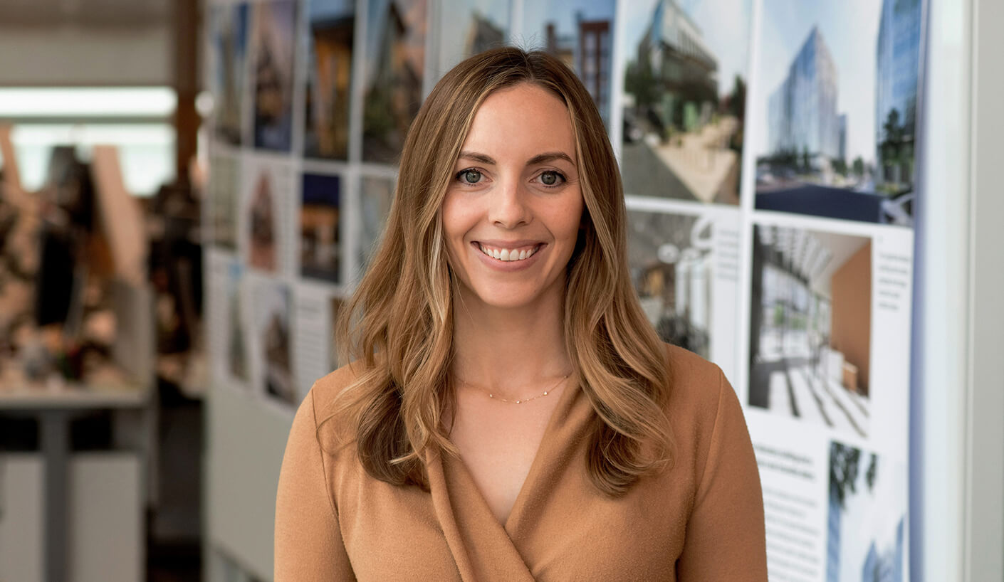 A woman with long hair smiles in front of architectural design boards in an office setting, wearing a tan sweater.
