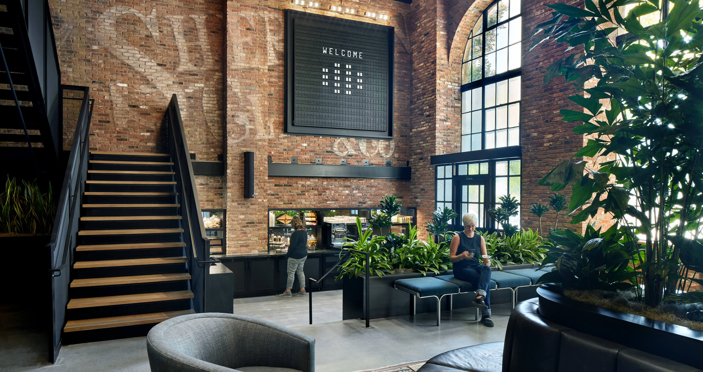 Interior view of a spacious lobby featuring brick walls, large windows, plants, a coffee area, and seating, with two people engaged in activity.