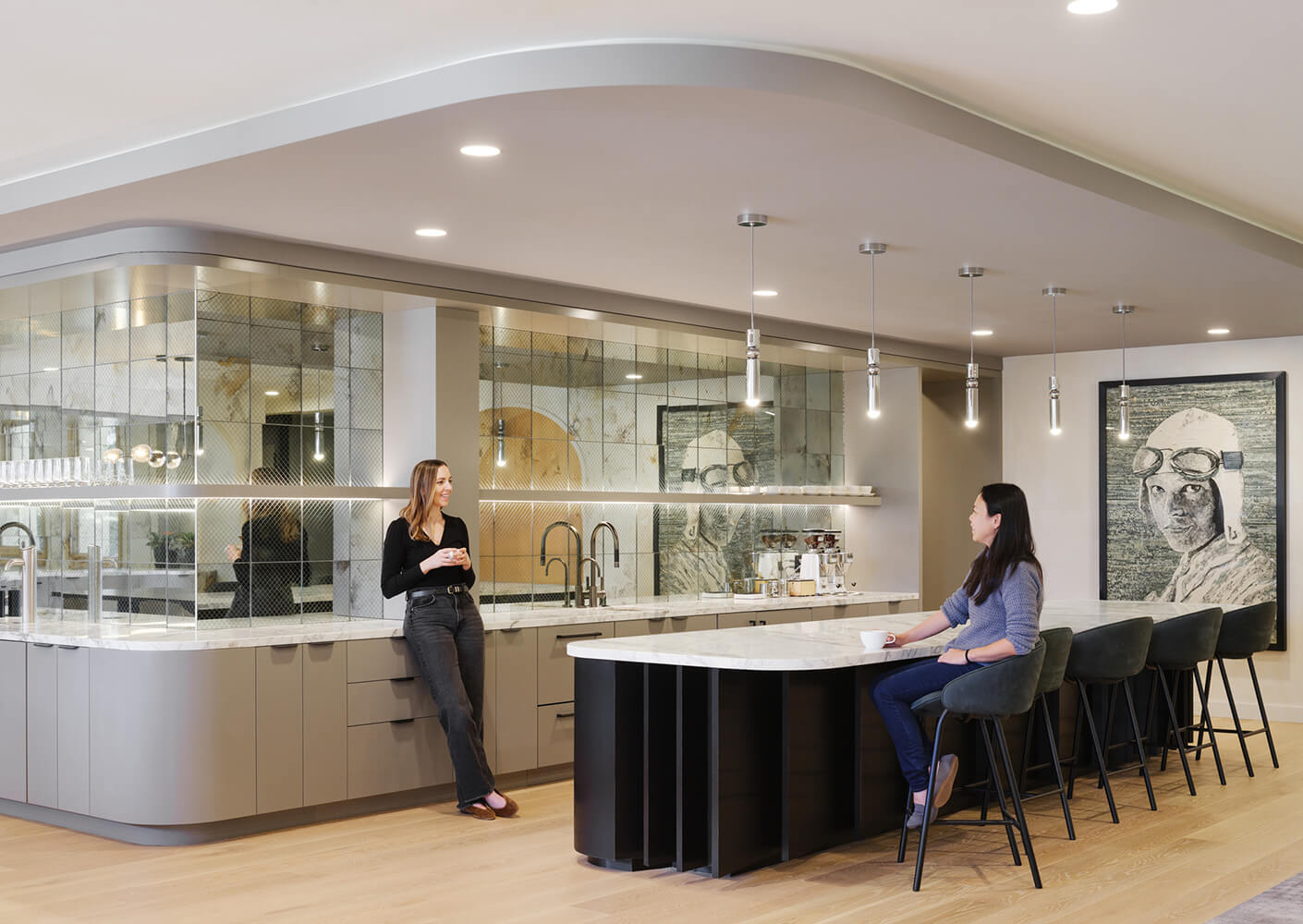 Two women converse in a sleek kitchen with a curved island, modern fixtures, and an art piece on the wall. Natural light enhances the space.