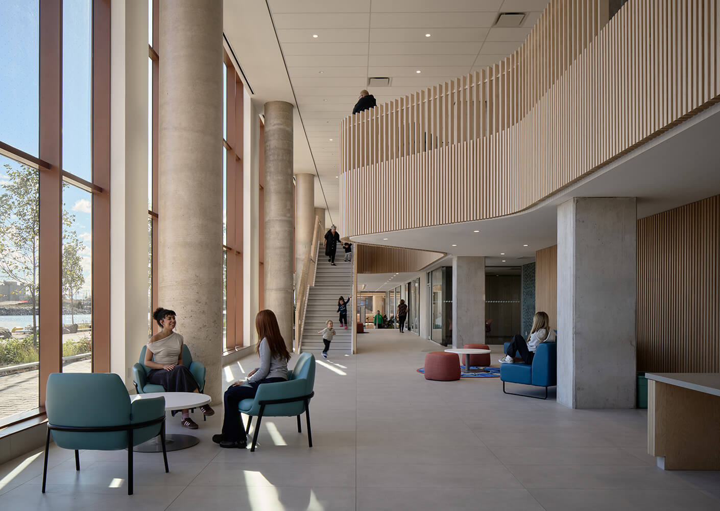 Bright interior space featuring large windows, seating areas, and a staircase, with people engaging and moving throughout the area.