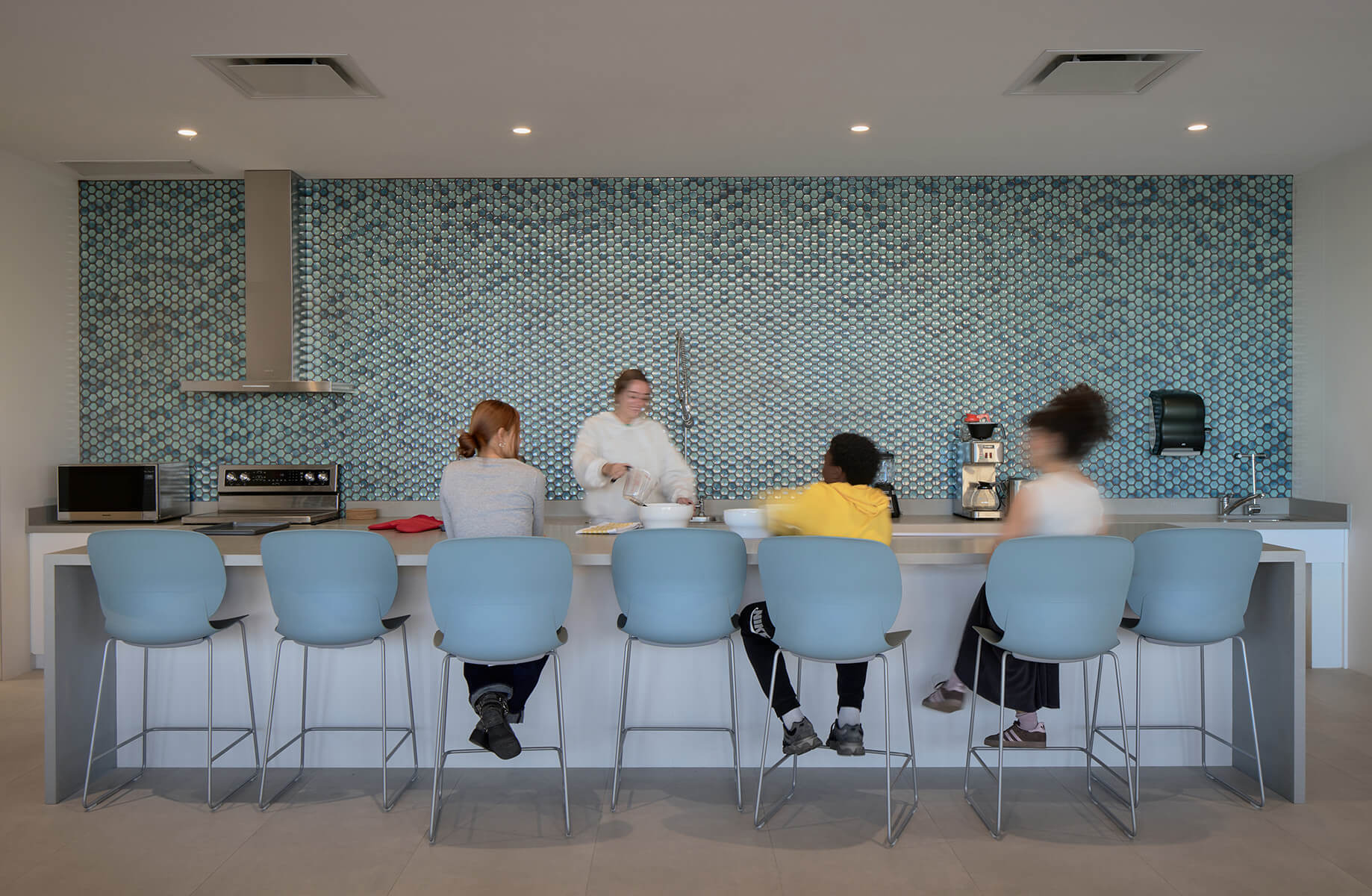 Group of four people at a kitchen island with blue stools, a tiled wall, and modern appliances, engaged in cooking and conversation.