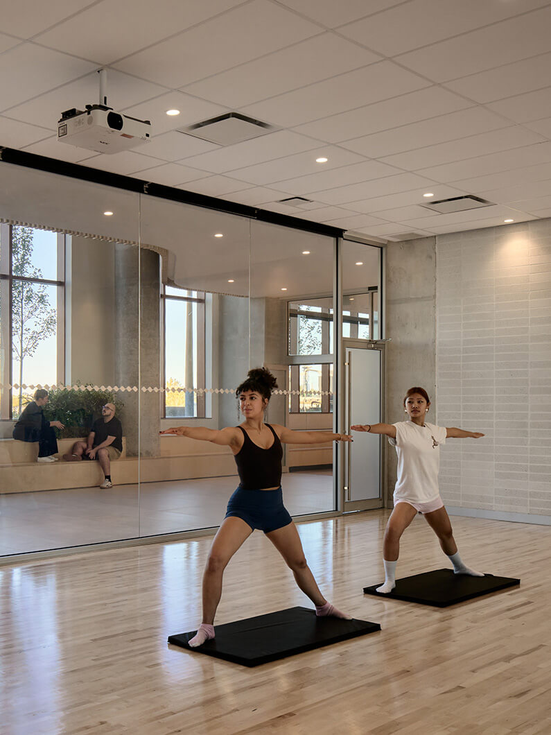 Two individuals practice yoga poses in a well-lit studio with mirrors and plants. A projector is mounted on the ceiling. People chat in the background.