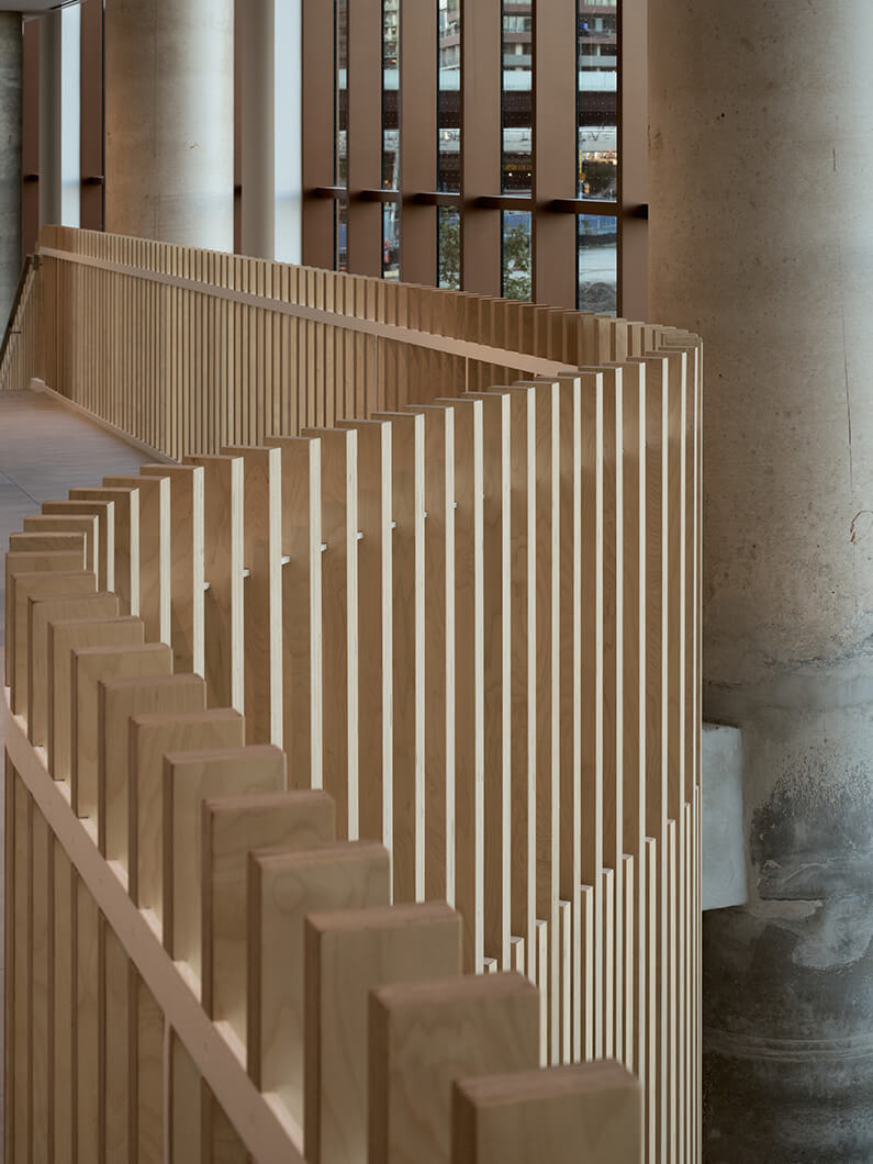 Wooden railing design in an interior space, featuring vertical slats and concrete columns with large windows in the background.