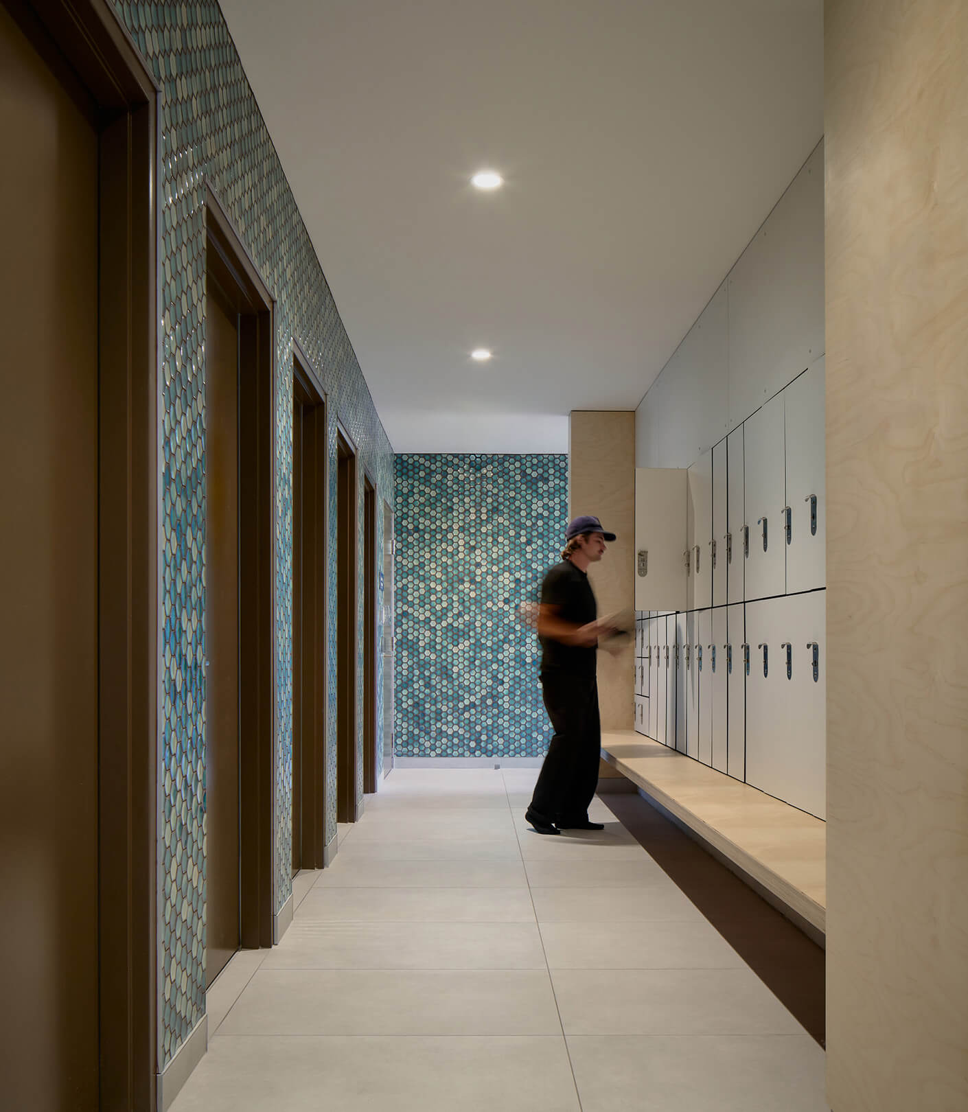 Interior view of a hallway with lockers and a person in a cap. Hexagonal tile wall adds texture to the space, with doors lining one side.