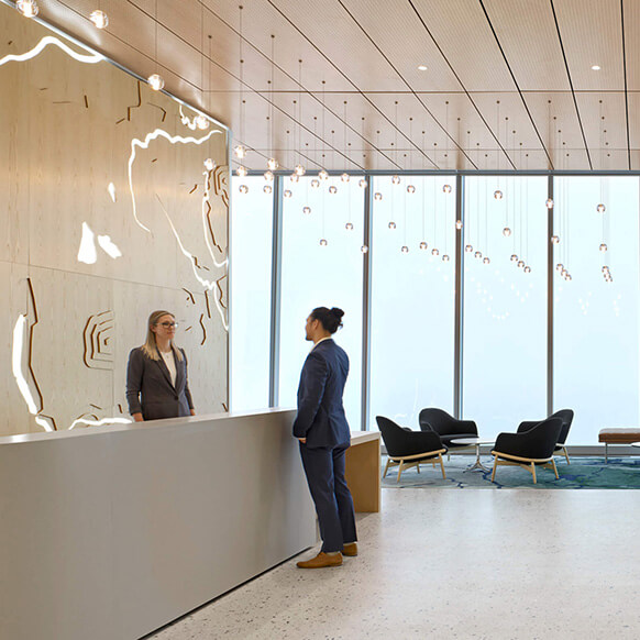 Office reception area with two people conversing at a sleek front desk, featuring wooden wall design and large windows with natural light.