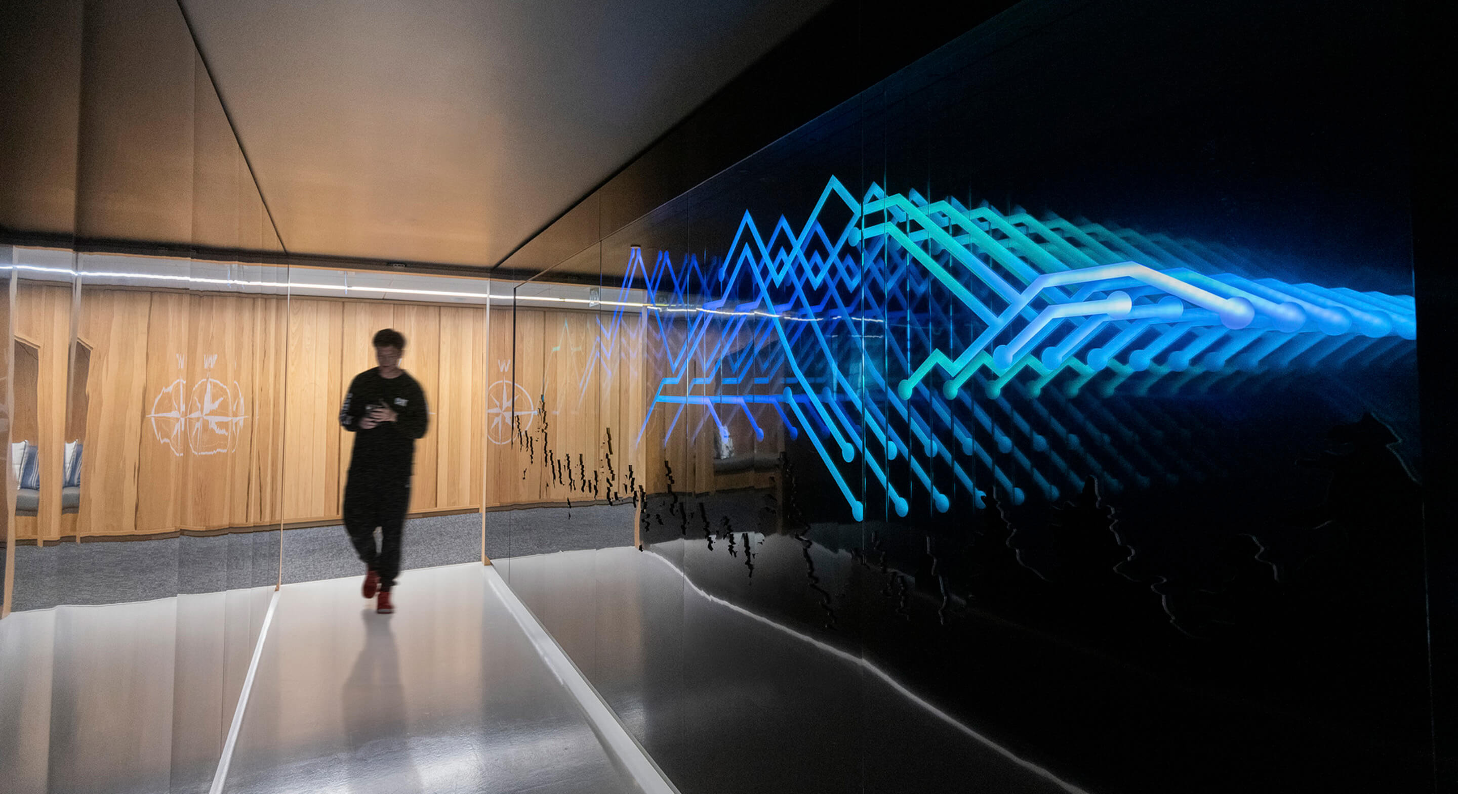 Person walking in a sleek hallway with a reflective surface and blue light patterns on a glass wall, featuring wooden paneling in the background.