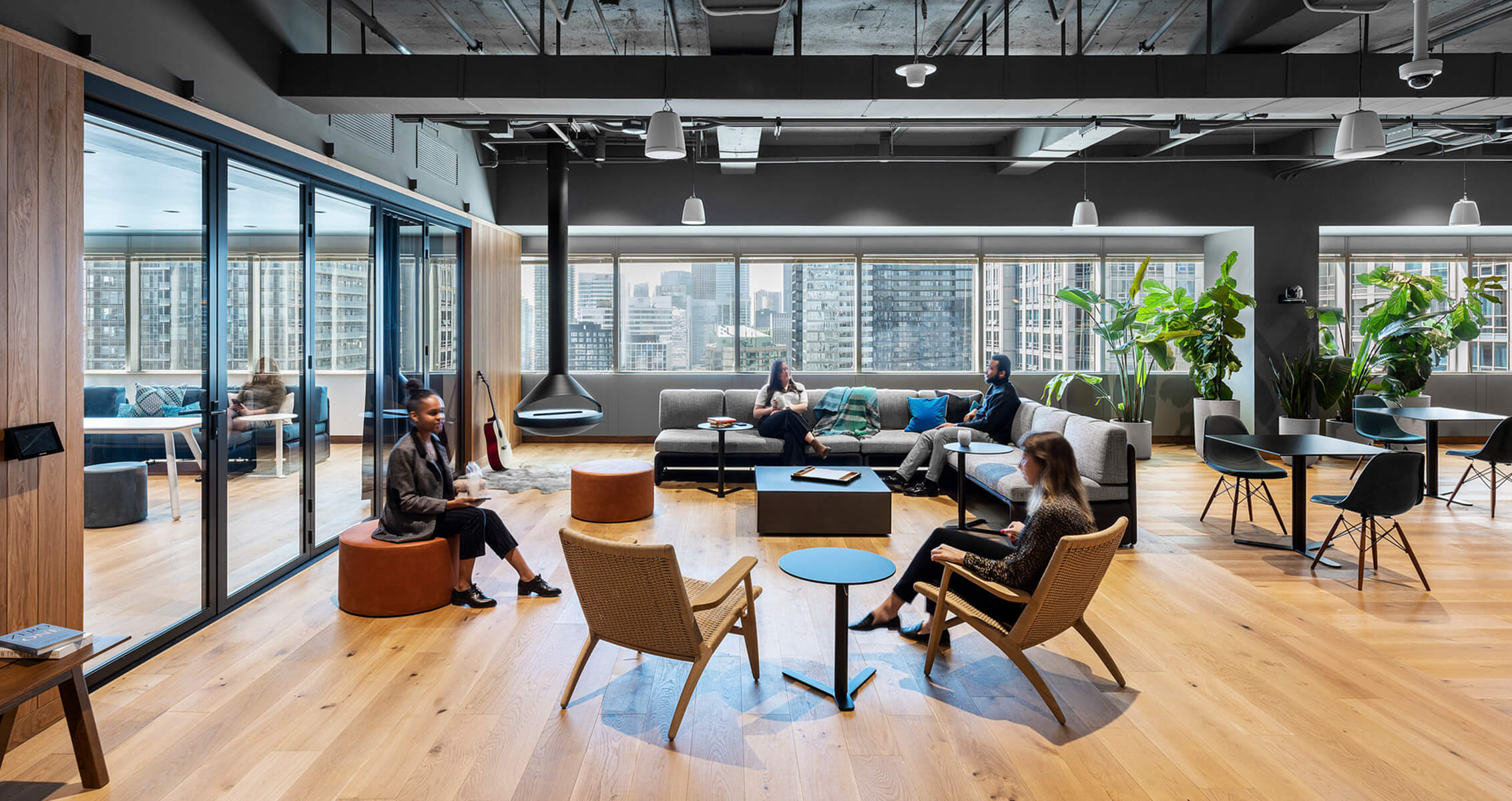 Cozy office lounge area with seating, large windows, greenery, and a mix of individuals engaged in conversation and relaxation.