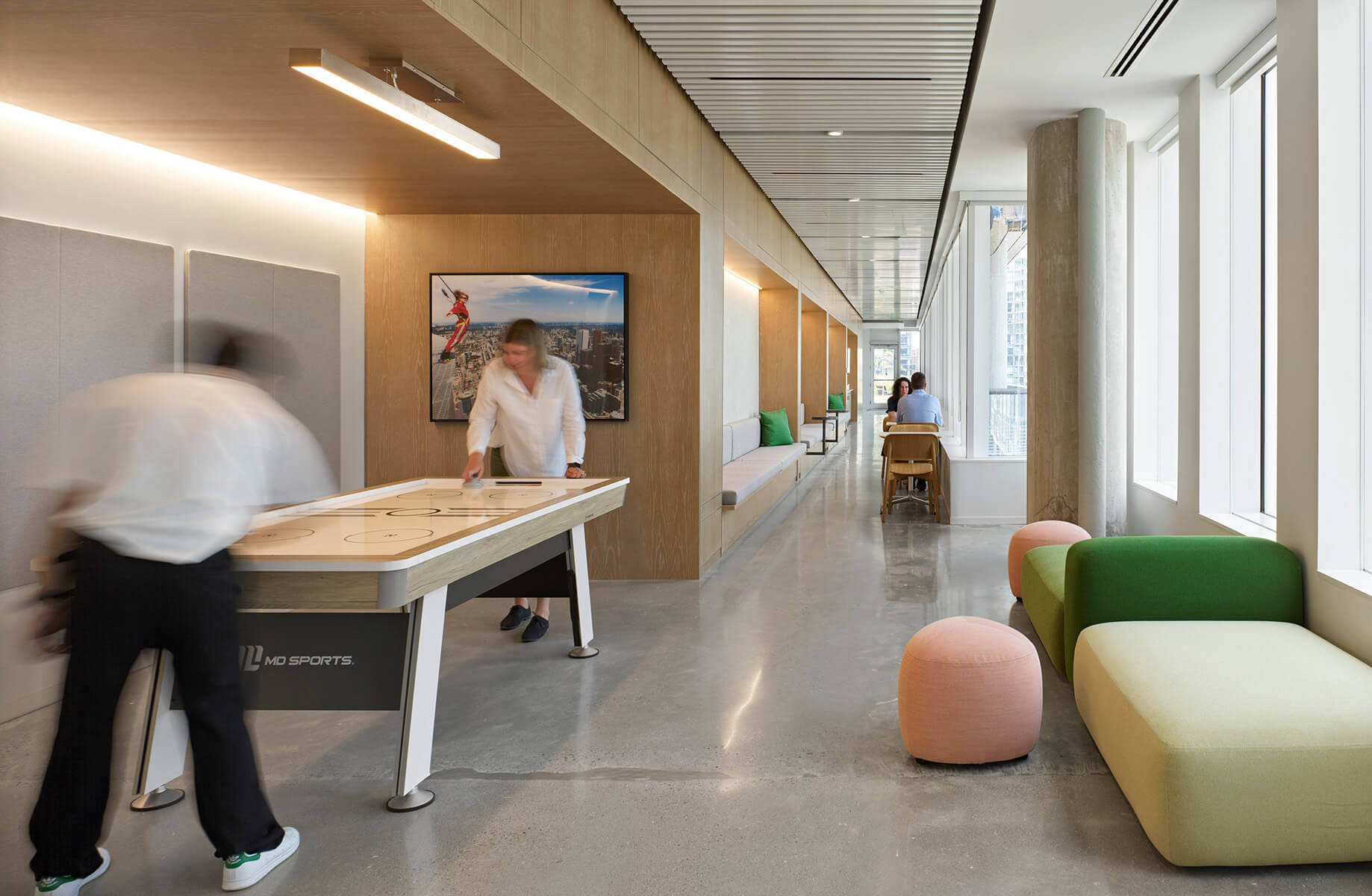 People engage in air hockey in a stylish hallway with colorful seating and windows, fostering a collaborative workspace environment.