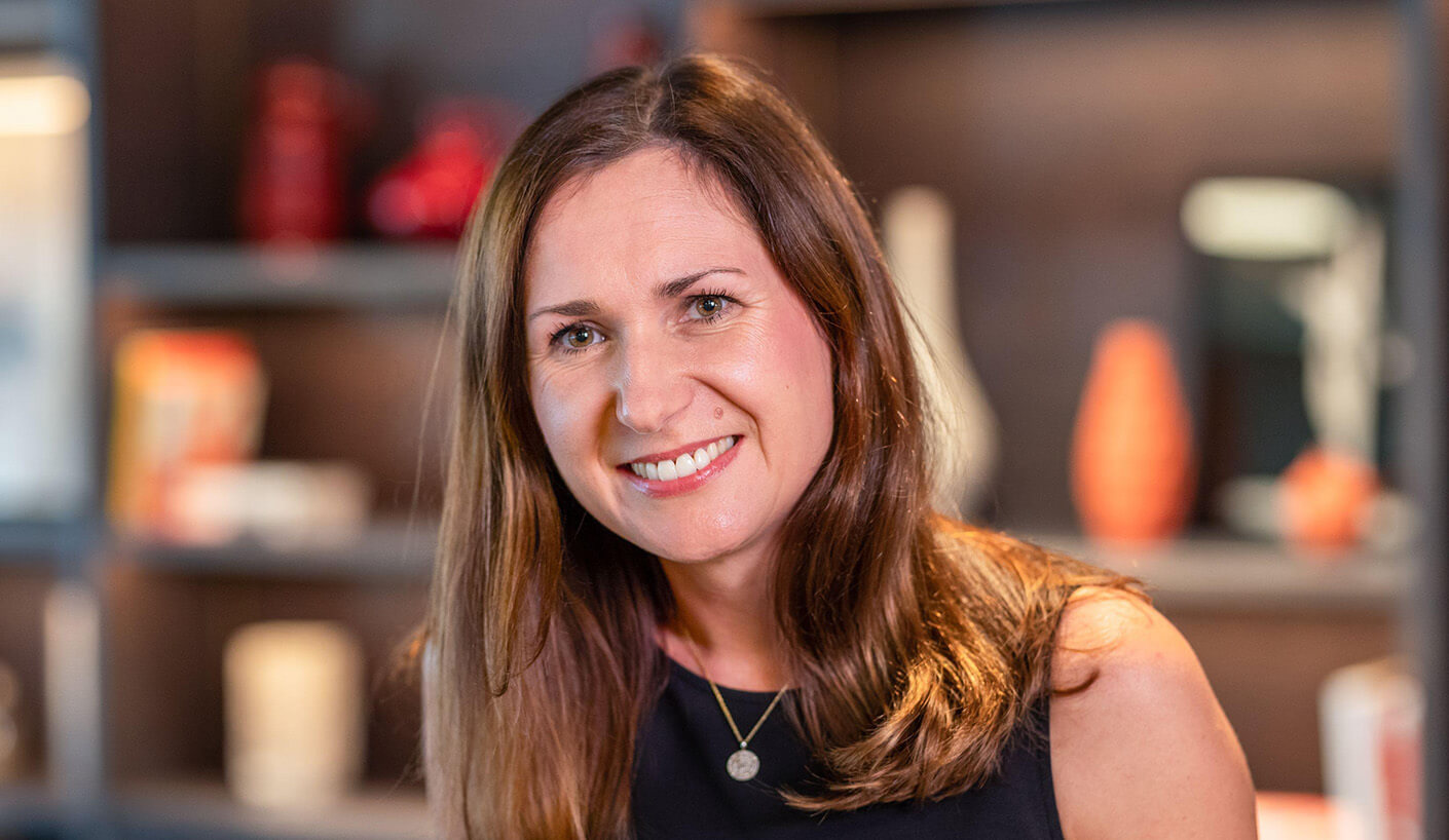 Smiling woman with long hair wearing a black top, posing in front of a shelf with decorative vases and various displayed items.