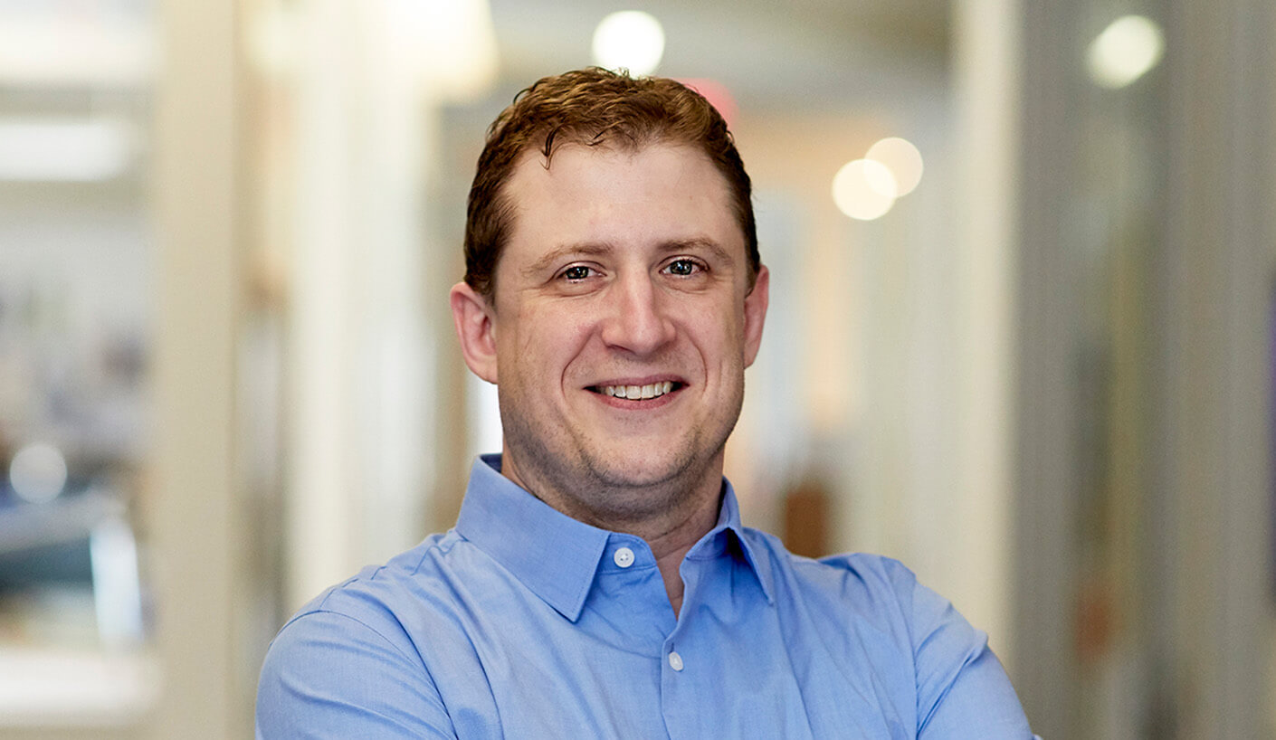 Smiling man in a light blue shirt stands with arms crossed in a well-lit hallway, featuring blurred background details of an office environment.