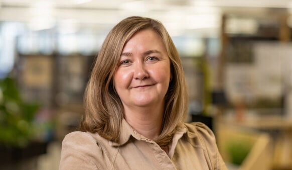 Professional woman with light brown hair smiling in an office setting, with a blurred background of plants and workstations.