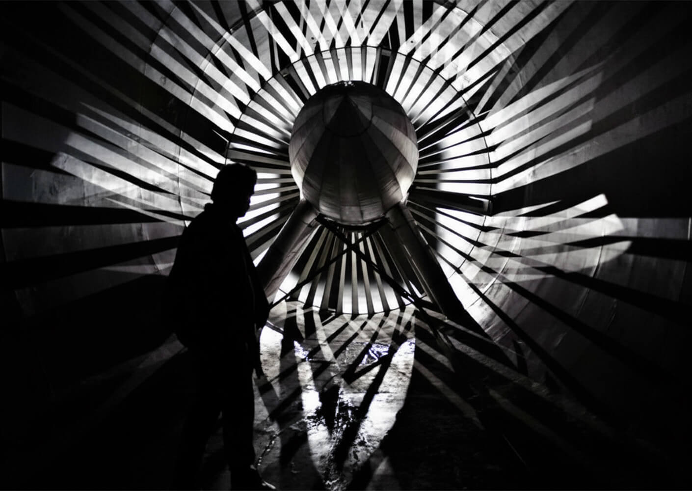 Silhouette of a person standing inside a large industrial turbine structure with dramatic light and shadow patterns on the walls.