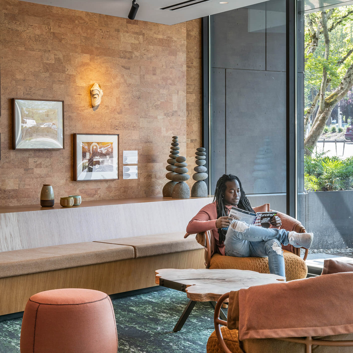 A person reads a magazine in a stylish lounge area with cork walls, art, and a cozy seating arrangement. Natural light filters through large windows.