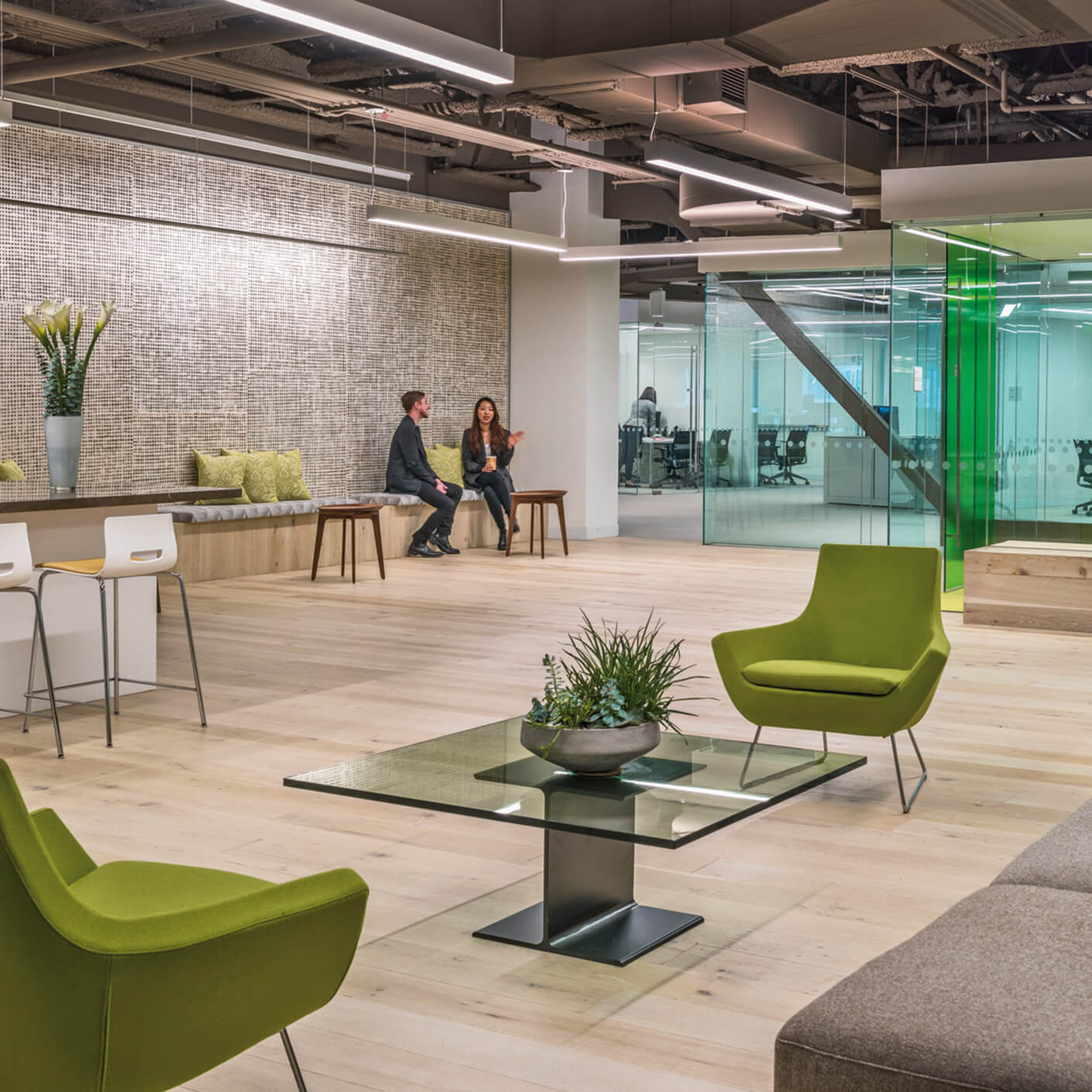 Two people converse in a spacious office lounge with green chairs, a glass table, and a textured wall. Natural light illuminates the area.