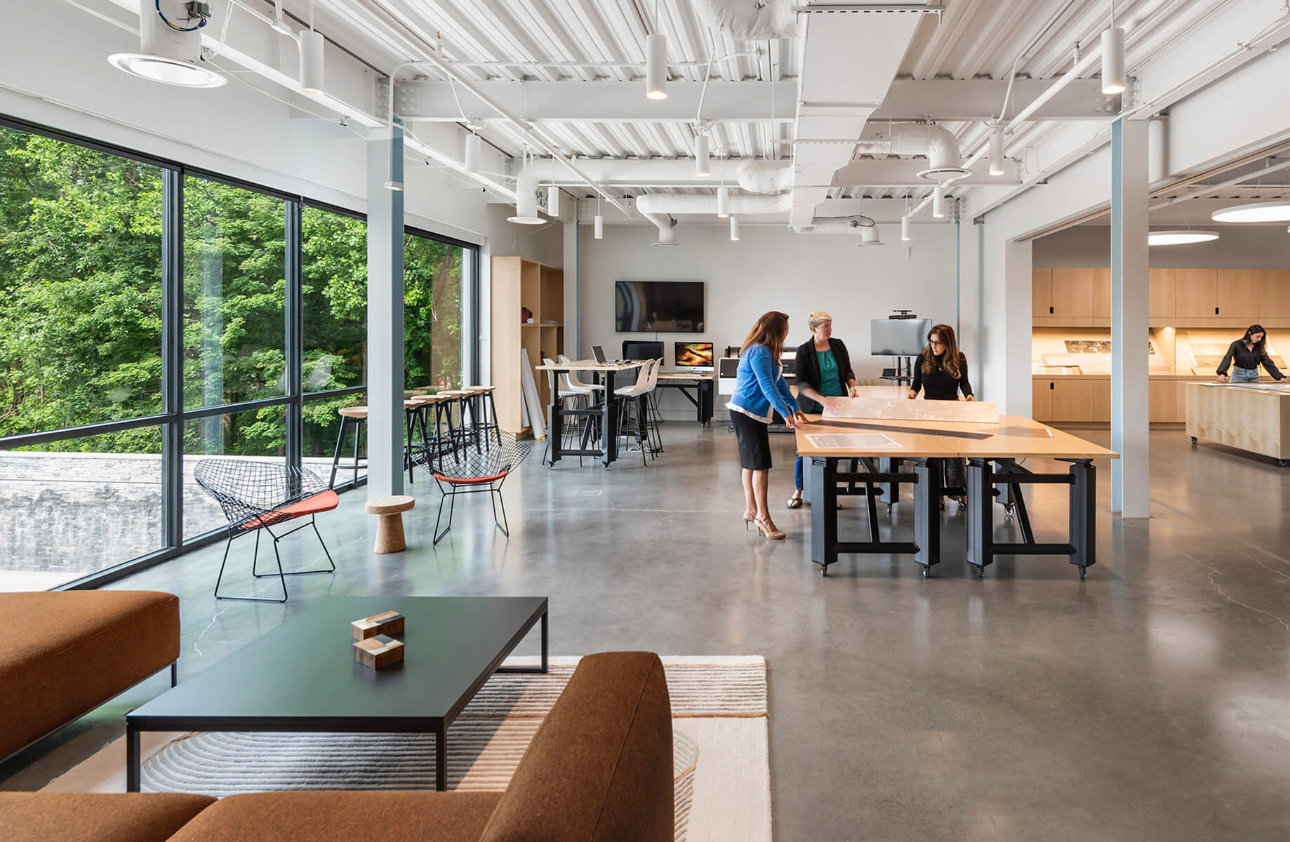 Four women collaborate around a large table in a bright, open workspace with large windows and modern furnishings. A kitchen area is visible.