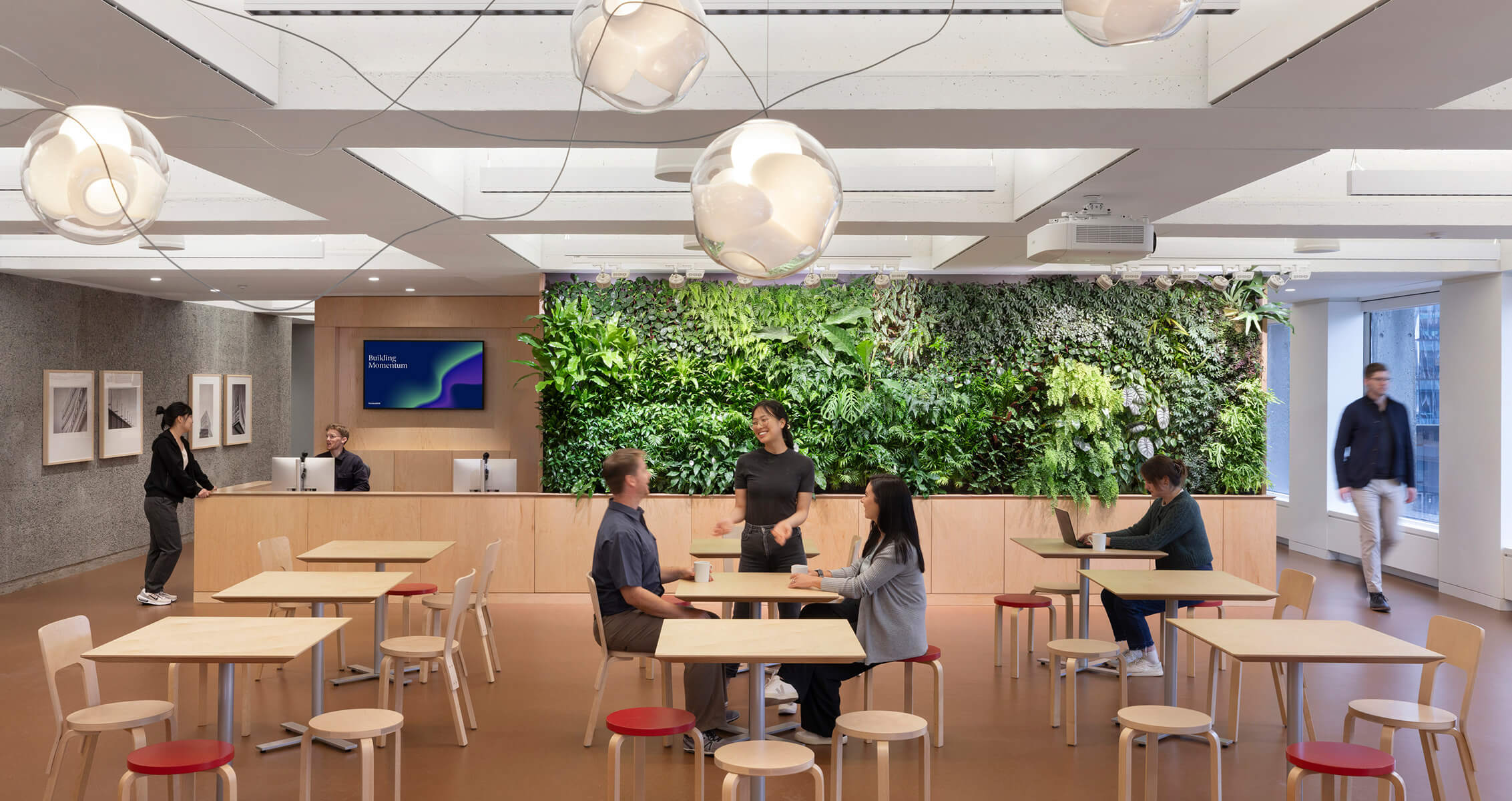 Interior view of a workspace featuring a reception area, greenery wall, and individuals engaged in conversation or working at tables.